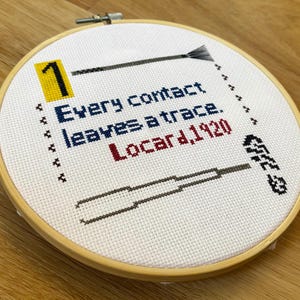 Puede incluir: Un bastidor de bordado circular de madera con tela blanca y texto bordado en punto de cruz que dice 1 Every contact leaves a trace. Locard, 1920. El diseño incluye un cuadrado amarillo, un pincel negro, una herramienta negra y símbolos.
