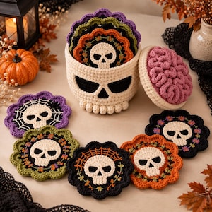May include: A set of round crochet coasters featuring skull designs in various colors including orange, green, purple, and black, stored in a matching cream-colored skull-shaped container with a pink brain-textured lid.