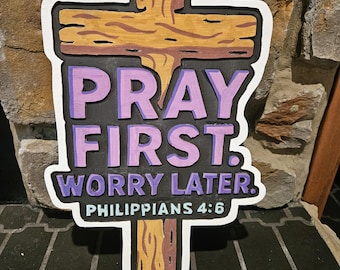 May include: Wooden cross-shaped sign with the text "PRAY FIRST. WORRY LATER. PHILIPPIANS 4:6". The cross has a light brown wooden grain design with a white border and black background. The text is in purple and white.
