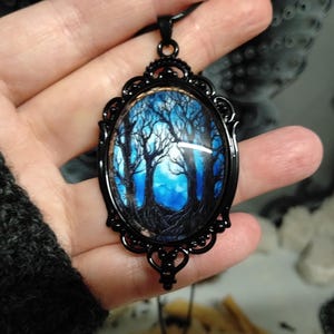 May include: Black oval pendant with a forest scene. The pendant has a detailed black filigree frame and a black cord. The image inside the pendant shows a blue and white forest scene with bare trees.