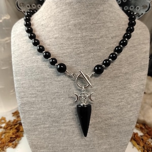 May include: A black beaded necklace with a silver toggle clasp. The pendant features a black, pointed stone with a silver triple moon and pentacle charm. The necklace is displayed on a grey mannequin.
