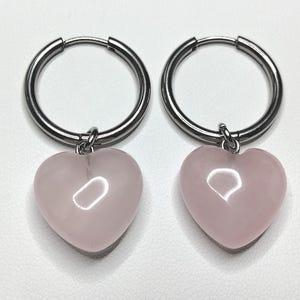 May include: A pair of silver-toned hoop earrings, each featuring a smooth, polished, light pink heart-shaped charm suspended from the bottom. The hoops are circular with a hinged closure mechanism, and the heart charms have a glossy finish.