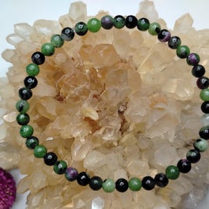 May include: A circular beaded bracelet featuring small, polished spherical stones in alternating shades of deep forest green, opaque black, and speckled magenta-pink, arranged in a continuous pattern on a thin, flexible cord.