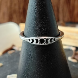 May include: A silver ring with a crescent moon design. The ring has a series of crescent moon cutouts across the top, creating a celestial pattern. The ring is displayed on a black stand, showcasing its details.