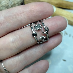 May include: A silver ring featuring celestial motifs: a sun, moon, and stars. The ring has a layered design and a small black stone. The ring is on a finger.