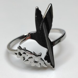 May include: A silver-toned metal ring featuring a cutout design of a hummingbird in flight positioned above two small, five-petalled flowers. The band is thin and polished, with the bird and floral elements integrated into the top section of the ring.