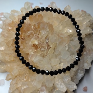 May include: A circular bracelet composed of small, faceted black beads arranged in a continuous loop. The beads have a reflective, polished surface that catches the light, creating a uniform appearance throughout the entire length of the jewellery piece.