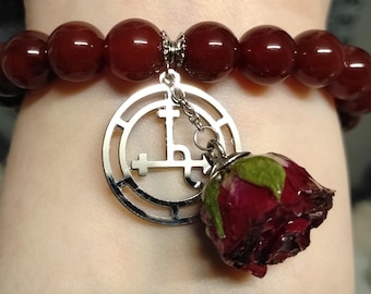 May include: A dark red beaded bracelet with a silver charm and a dried rose pendant. The charm features a symbol within a circle. The rose is a deep red color with green leaves.