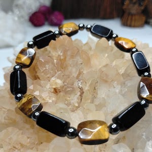 May include: A beaded bracelet featuring alternating rectangular tiger eye beads with golden-brown chatoyancy and smooth black onyx beads, separated by small, round, metallic-finish hematite spacer beads arranged in a circular pattern.
