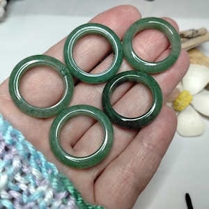May include: Five green jade rings of varying shades are displayed. The rings are smooth and polished, with a circular design. The rings are held in a hand, showcasing their natural colour and texture. The rings are a jewellery item.