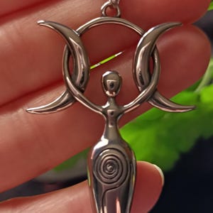 May include: A silver-toned pendant featuring a stylized depiction of a female figure with arms raised, encircled by a ring and crescent moons. The pendant has a spiral design on the torso and hangs from a matching chain.