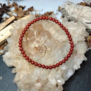 May include: A red beaded bracelet made of small, round beads. The bracelet is presented on a large, clear crystal formation. The background features dried leaves, sage, and wooden sticks.