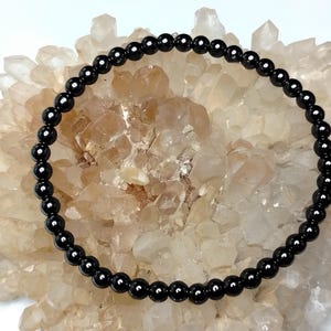 May include: A circular bracelet composed of small, uniform, polished black spherical beads strung together. The beads have a smooth, reflective surface and are arranged in a continuous loop, creating a simple and consistent design throughout the piece.