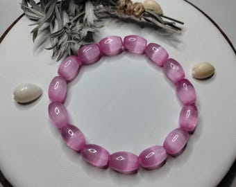 May include: A circular bracelet composed of smooth, barrel-shaped beads in a translucent pink hue. Each bead features a distinct chatoyant band that reflects light, creating a shimmering effect. The beads are strung together in a continuous loop.