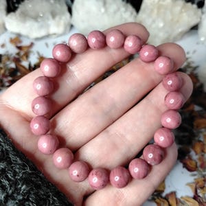 May include: A pink rhodonite beaded bracelet. The bracelet is composed of round, polished beads in varying shades of pink. The bracelet is held in a hand, with a dark gray sleeve visible in the corner. Crystals and dried flowers are in the background.