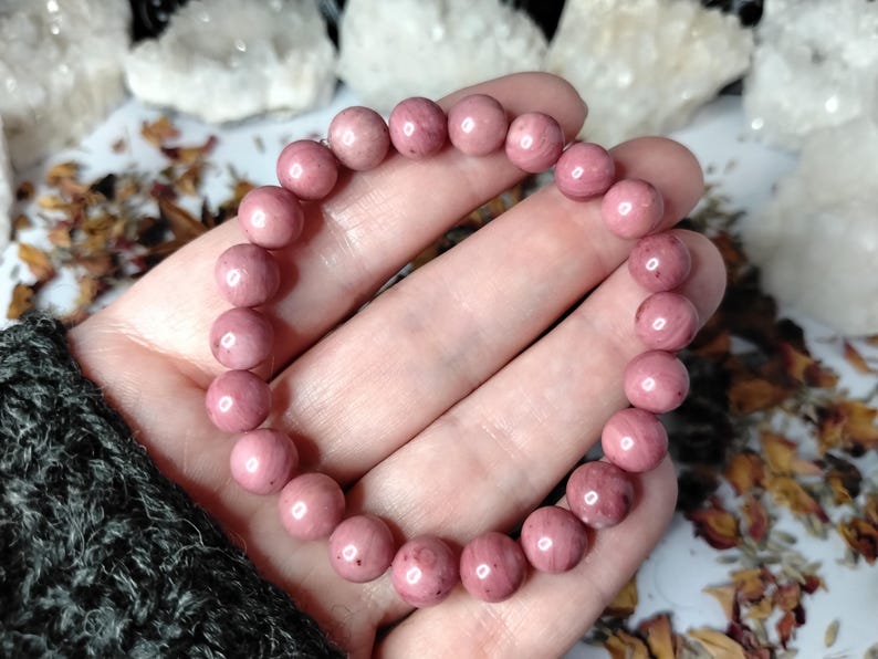 May include: A pink rhodonite beaded bracelet. The bracelet is composed of round, polished beads in varying shades of pink. The bracelet is held in a hand, with a dark gray sleeve visible in the corner. Crystals and dried flowers are in the background.