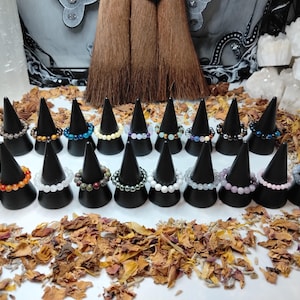 May include: A selection of beaded rings in various colours, displayed on black cone-shaped stands. The rings feature beads in shades of orange, white, blue, yellow, and purple. The background includes dried leaves and decorative elements.