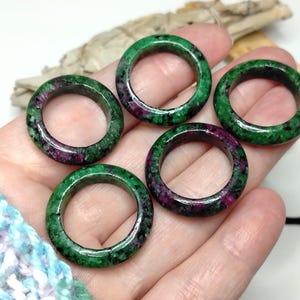 May include: Five gemstone rings in green and red. The rings are made of a polished stone material, displaying a speckled pattern. The rings are round and appear to be of a medium size. The rings are likely to be around 2 cm in diameter.