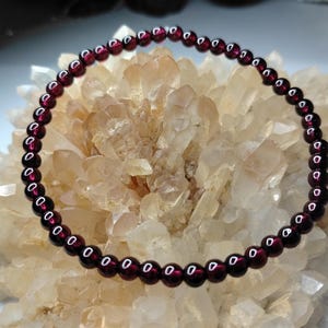 May include: A bracelet made of small, round, deep red garnet beads. The beads are strung together to form a complete circle. The gemstones are a rich, dark red colour. The bracelet is displayed on a light-coloured, crystalline surface.