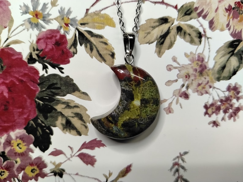 May include: A crescent moon pendant necklace with a silver chain. The moon-shaped pendant features a mix of colours, including red, green, and black. The necklace is displayed against a floral background.