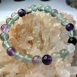 May include: A beaded bracelet composed of smooth, spherical fluorite gemstones. The beads feature a translucent appearance with a colour palette alternating between shades of deep purple, pale green, and clear white, strung together on a flexible cord.