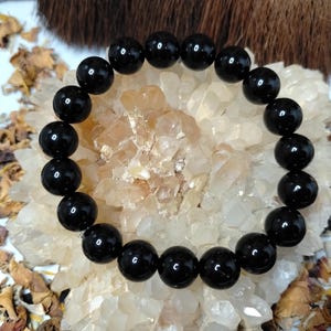 May include: A black beaded bracelet featuring smooth, spherical beads. The bracelet is presented on a bed of crystals and dried botanicals. The beads are uniform in size and strung together, forming a simple, elegant accessory.