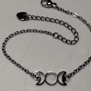 May include: A silver-toned metal chain bracelet featuring a central charm design of a full moon flanked by two crescent moons. The bracelet includes a delicate link chain, an adjustable extender, a lobster claw clasp, and a small heart-shaped charm.