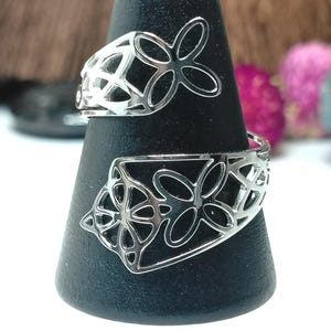 May include: A silver-toned bypass ring featuring an openwork design with repeating four-petaled floral or butterfly-like motifs. The metal band wraps around the finger, showcasing intricate cutouts that create a delicate, airy appearance.