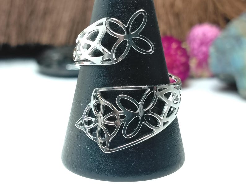 May include: A silver-toned bypass ring featuring an openwork design with repeating four-petaled floral or butterfly-like motifs. The metal band wraps around the finger, showcasing intricate cutouts that create a delicate, airy appearance.