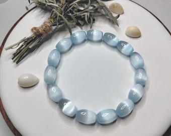 May include: A stretch bracelet composed of smooth, oval-shaped light blue cat's eye beads. The beads exhibit a chatoyant effect, creating a shimmering band of light across their surfaces. The bracelet is arranged in a circular shape on a white surface.