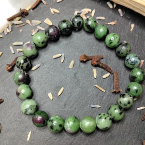 May include: A green and maroon gemstone bead bracelet. The bracelet is made of round beads and is displayed on a dark surface, with cloves and grains scattered around. The beads have a speckled pattern.