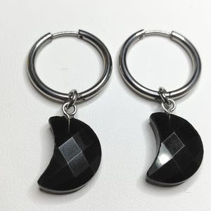 May include: A pair of silver-toned hoop earrings featuring dangling black crescent moon charms. Each charm has a faceted, geometric surface texture. The hoops are circular with a hinged closure mechanism, and the charms are attached via small silver rings.