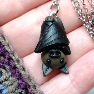 May include: A necklace featuring a sculpted black bat pendant hanging upside down with folded wings. The bat has a dark grey face, small black eyes, and pointed ears. It is attached to a silver-toned metal chain with a lobster claw clasp.