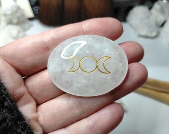 Puede incluir: Una piedra blanca translúcida de forma ovalada con una superficie lisa y pulida. Centrado en la piedra hay un símbolo grabado de color dorado que presenta un círculo central flanqueado por dos lunas crecientes, el motivo de la triple luna.