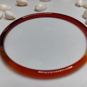 May include: A smooth, circular bangle bracelet crafted from translucent material with a gradient of deep red to amber orange tones, surrounded by several small, off-white, oval-shaped seashells arranged in a scattered pattern.