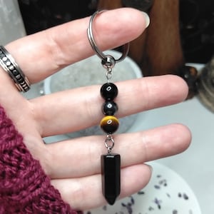 May include: A keyring featuring a silver-toned metal ring attached to a vertical arrangement of beads and a stone pendant. The design includes a smooth black bead, a metallic grey bead, a golden-brown tiger's eye bead, and a polished black stone point.