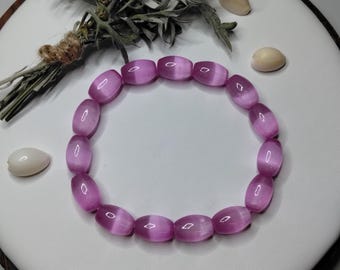 May include: A circular bracelet composed of smooth, translucent, light purple barrel-shaped beads. Each bead features a distinct chatoyant band running through the center, creating a shimmering effect. The beads are strung together on an elastic cord.