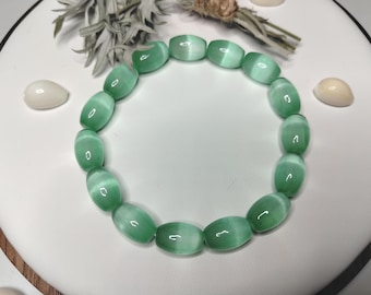 May include: A stretch bracelet composed of smooth, barrel-shaped beads in a translucent light green hue. Each bead features a distinct chatoyant band that creates a shimmering cat's eye effect, reflecting light across the surface of the stones.