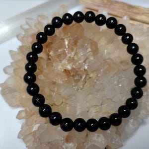 May include: A circular bracelet composed of smooth, polished black spherical beads strung together. The beads have a glossy finish that reflects light, creating small white highlights on their surfaces. The bracelet is arranged in a ring shape.