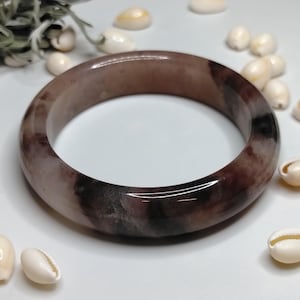 May include: A smooth, polished bangle bracelet featuring a marbled pattern with deep brown, black, and translucent mauve tones. The circular band has a glossy finish and a rounded profile, showcasing natural colour variations throughout the stone material.