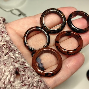 May include: Five circular rings crafted from mahogany obsidian, each featuring a smooth, polished surface with distinct dark brown and black mottled patterns. The rings are arranged in a cluster, showcasing their rounded band design and natural stone texture.