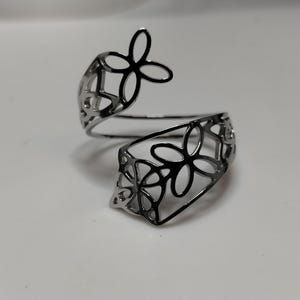 May include: A silver-toned bypass ring featuring an openwork design of interconnected four-petaled flower shapes. The metal band is polished and reflective, with a continuous cutout pattern that wraps around the finger in a spiral formation.
