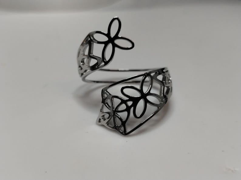 May include: A silver-toned bypass ring featuring an openwork design of interconnected four-petaled flower shapes. The metal band is polished and reflective, with a continuous cutout pattern that wraps around the finger in a spiral formation.
