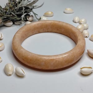 May include: A smooth, round bangle bracelet in a soft peach-toned, mottled stone material. The bracelet has a thick, polished band with a consistent circular shape and a glossy finish, displayed alongside small white seashells and a dried herb bundle.