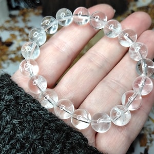 May include: A clear quartz crystal bracelet with round beads. The beads are strung together on a thin, elastic cord. The bracelet is held in a hand, with a dark grey knitted sleeve visible in the corner. The beads are approximately 1.3 cm in diameter.