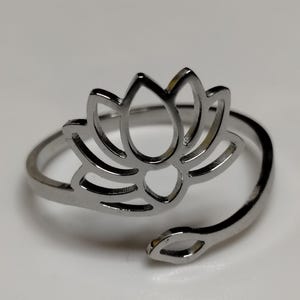 May include: A silver-toned metal ring featuring an openwork lotus flower design. The band is adjustable, with one end terminating in the lotus shape and the other end featuring a small, leaf-shaped accent. The surface has a polished, reflective finish.