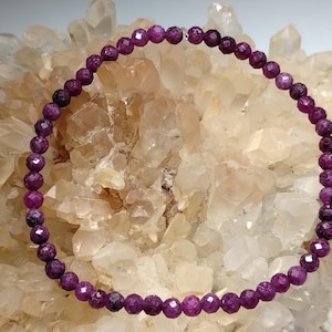 May include: A circular strand of small, faceted, deep magenta-coloured beads arranged in a ring. The beads have a textured, reflective surface and are uniform in size, creating a continuous loop of dark pink gemstone-like spheres.