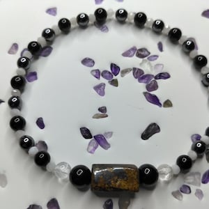 May include: A beaded bracelet featuring a repeating pattern of glossy black spherical beads and smaller translucent white beads. A single rectangular, textured brown and metallic-flecked bead is positioned as a central focal point on the strand.