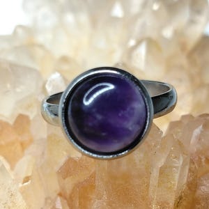 May include: A silver ring with a large, round, polished purple gemstone. The gemstone is set in a bezel setting, and the ring band is polished silver. The ring is displayed against a backdrop of light-coloured crystals.