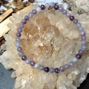May include: A bracelet made of small, round, lavender-coloured beads. The bracelet is presented on a cluster of light-coloured crystals, with other natural elements in the background. The beads have a smooth, polished appearance.